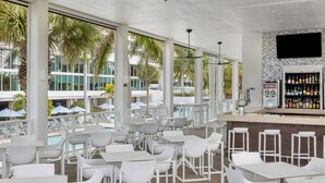 Un área de bar moderna al aire libre con mobiliario blanco y grandes ventanales con vistas a una piscina y palmeras.