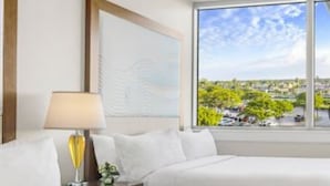 Habitación de hotel con dos camas, un ventilador de techo, una ventana con vista a la ciudad y una lámpara sobre una mesita de noche.