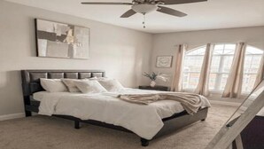 Une chambre à coucher avec un lit, un ventilateur de plafond, une fenêtre avec des rideaux et un tableau au mur.