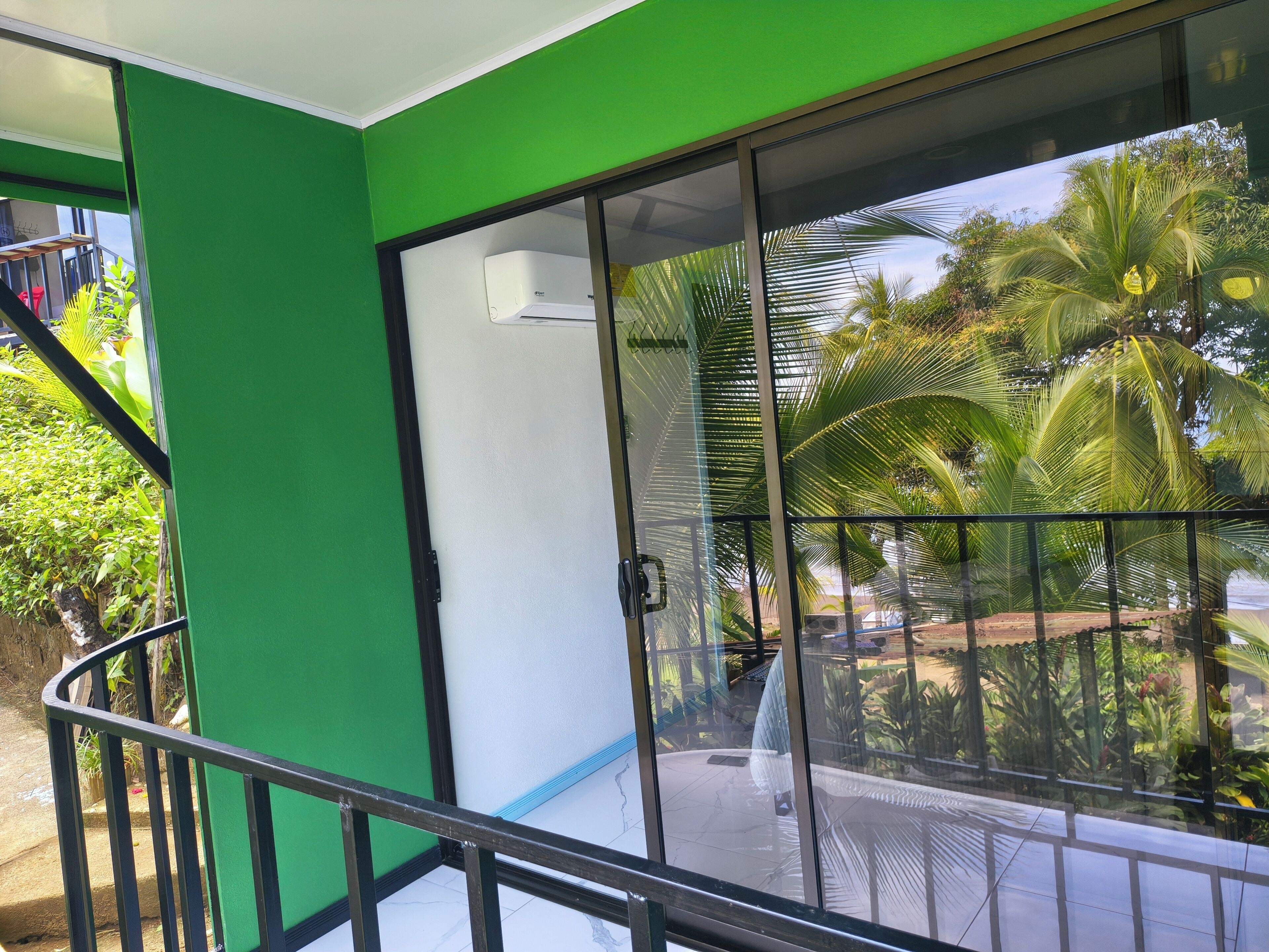 A room with a sliding glass door leading to a balcony with a view of palm trees and a garden.
