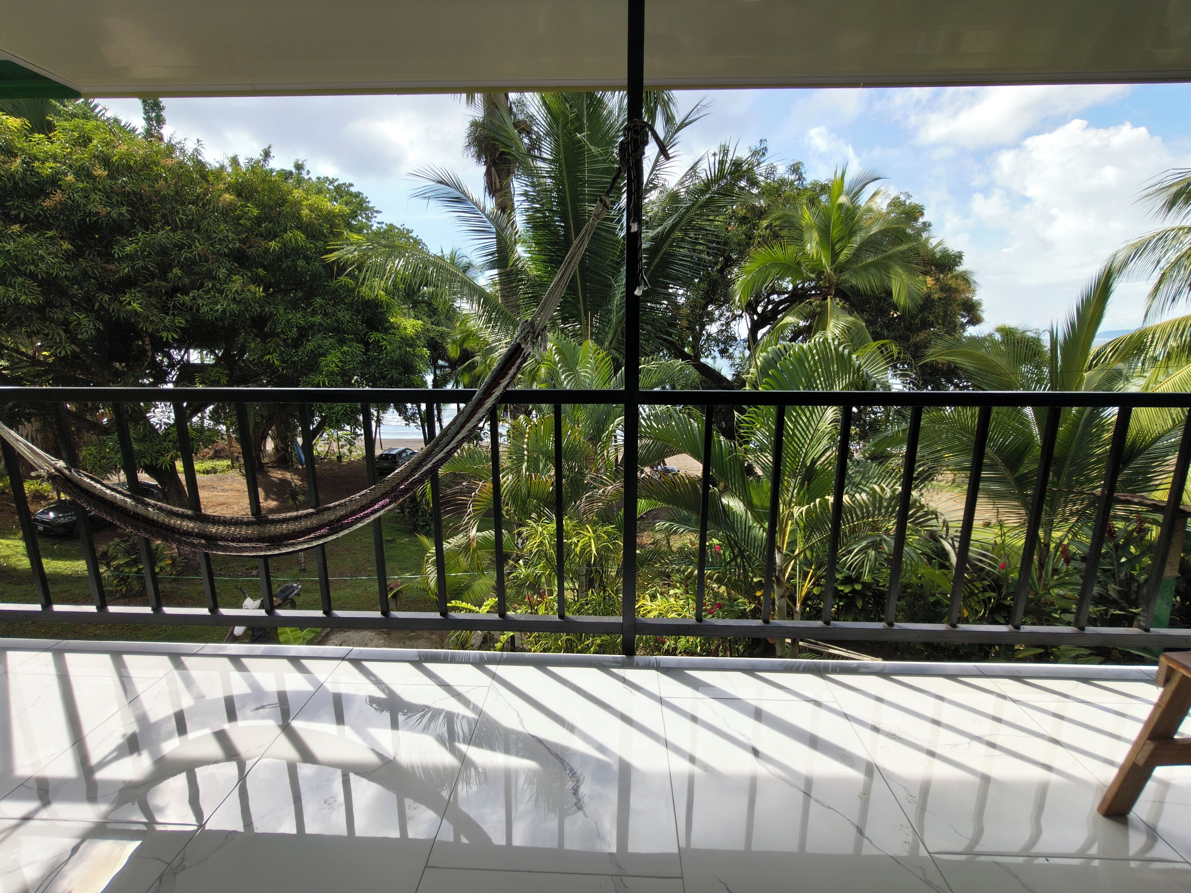 A balcony with a hammock, a tiled floor, and a view of palm trees and a beach.