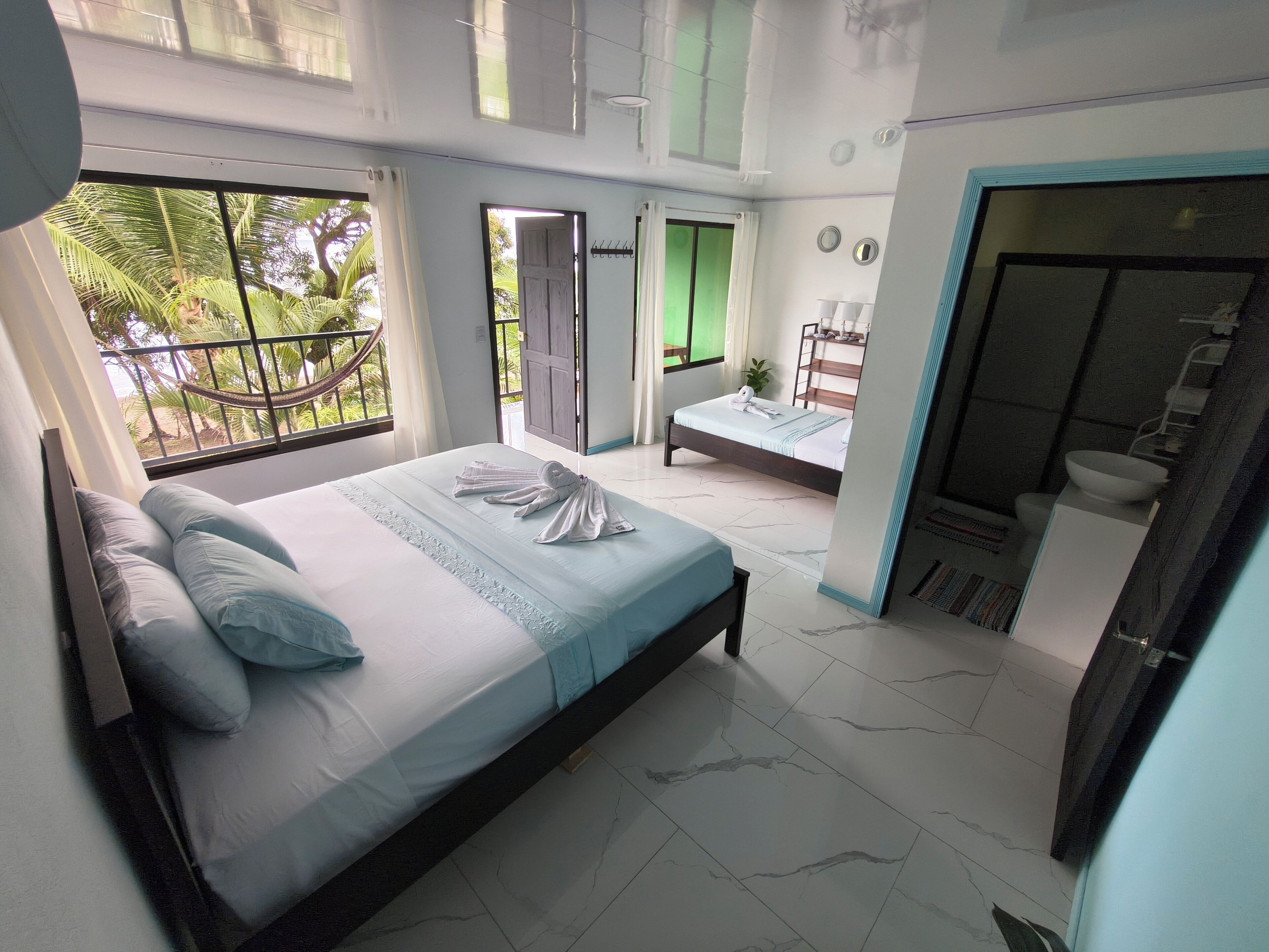 A bedroom with a bed, a small bedside table, a window with a view of palm trees, and a door leading to another room.