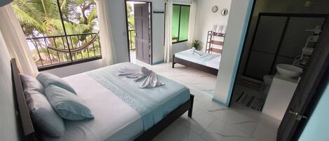A bedroom with a bed, a small bedside table, a window with a view of palm trees, and a door leading to another room.