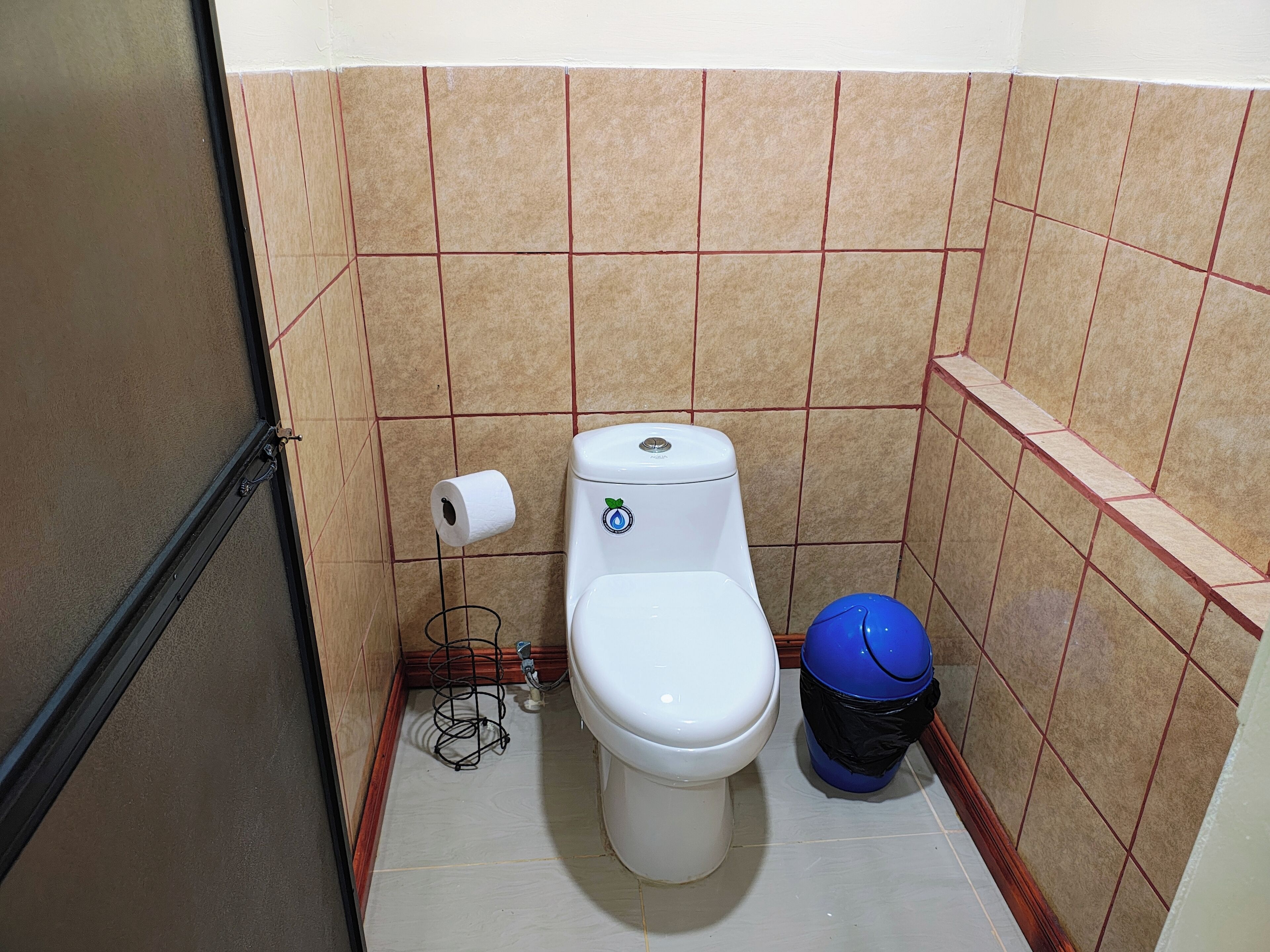 Une petite salle de bain avec des toilettes, un rouleau de papier toilette, une poubelle bleue et un étagère en métal avec un tuyau.