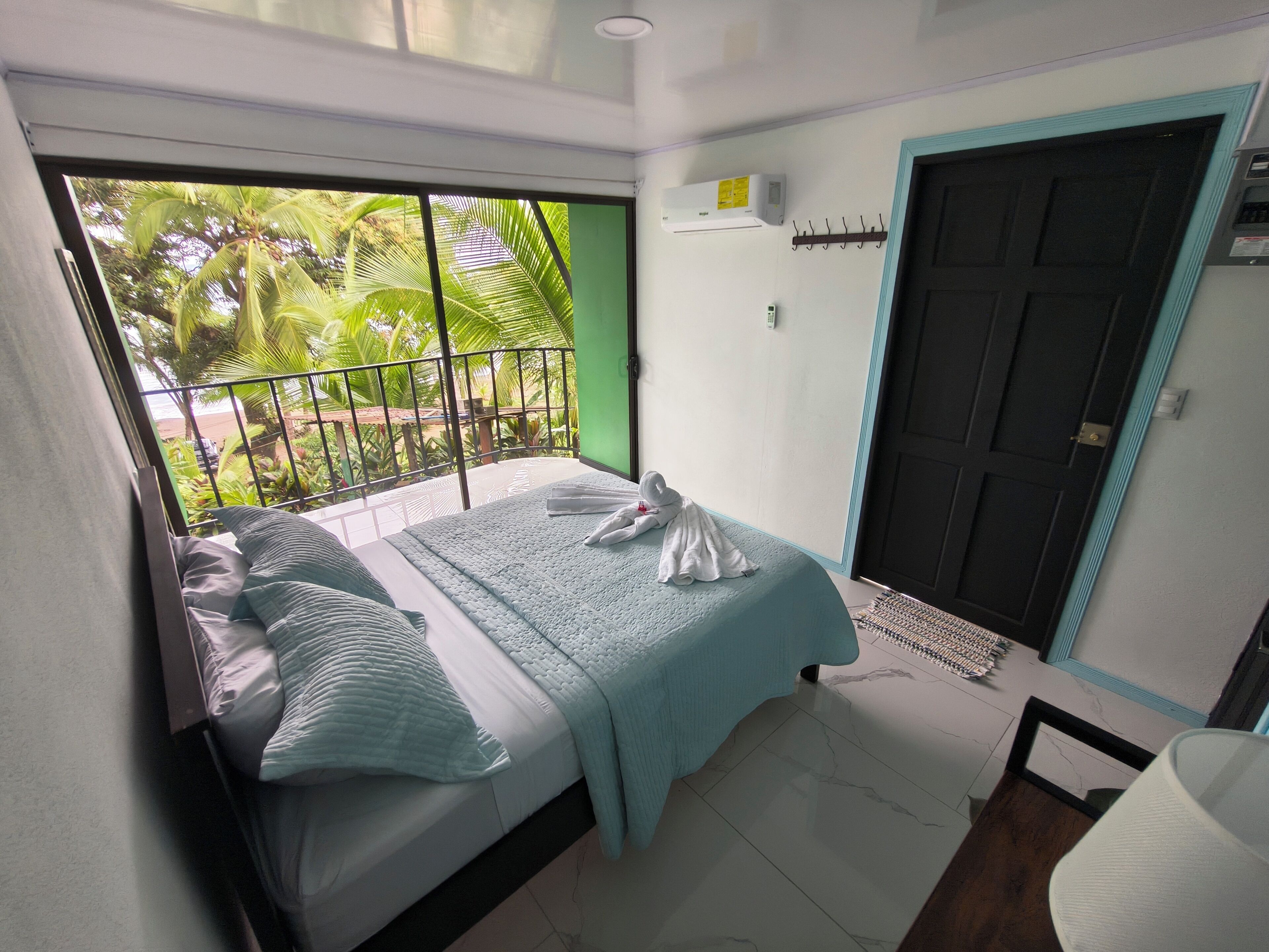 Quarto com cama, janela com vista para vegetação, porta e ar condicionado.