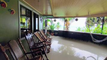 A spacious patio with wooden chairs and a dining table, surrounded by potted plants and a hammock.
