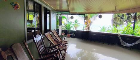 A spacious patio with wooden chairs and a dining table, surrounded by potted plants and a hammock.