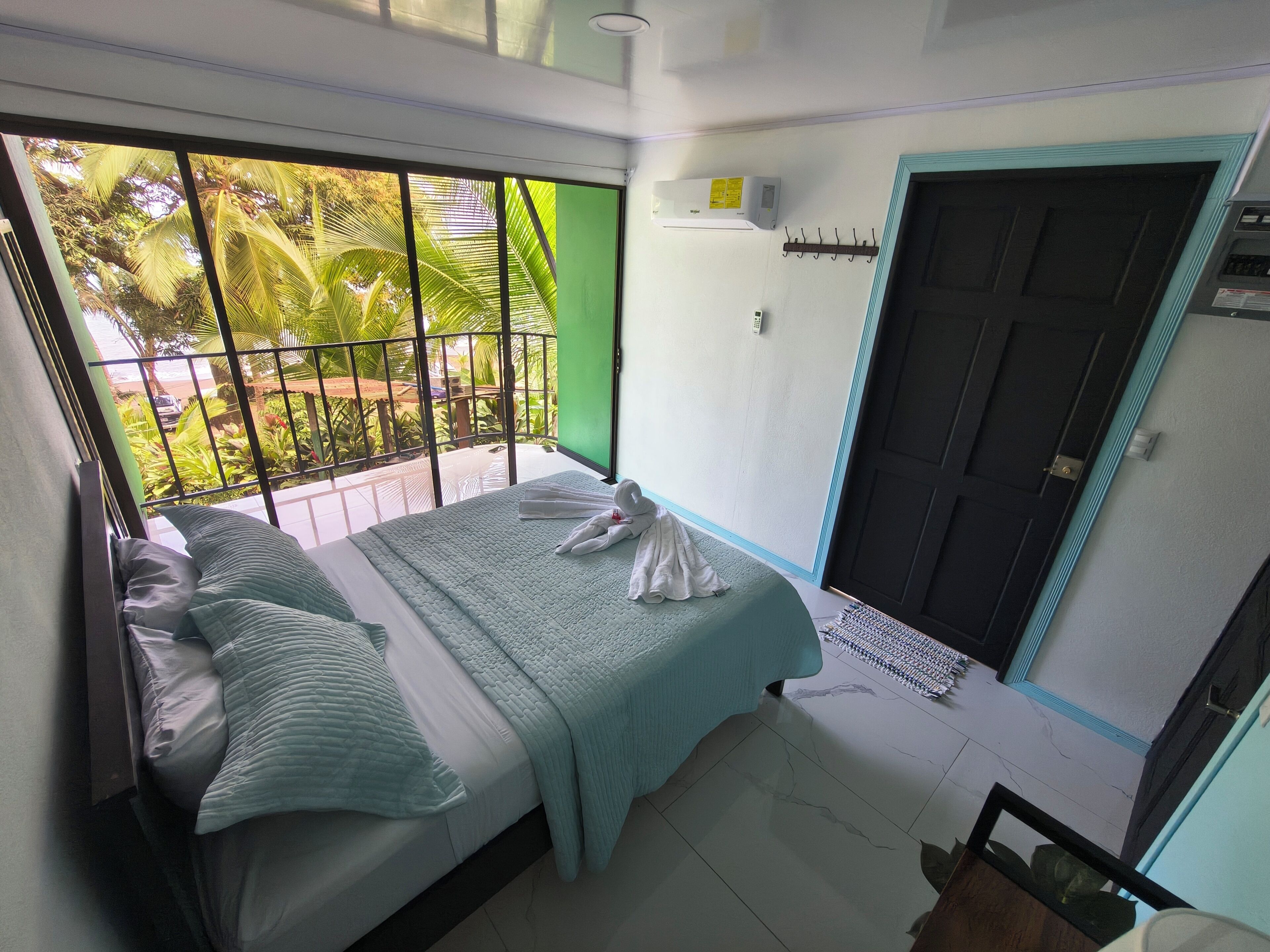 A room with a bed, a window with a view of palm trees, a door, and a wall-mounted air conditioner.