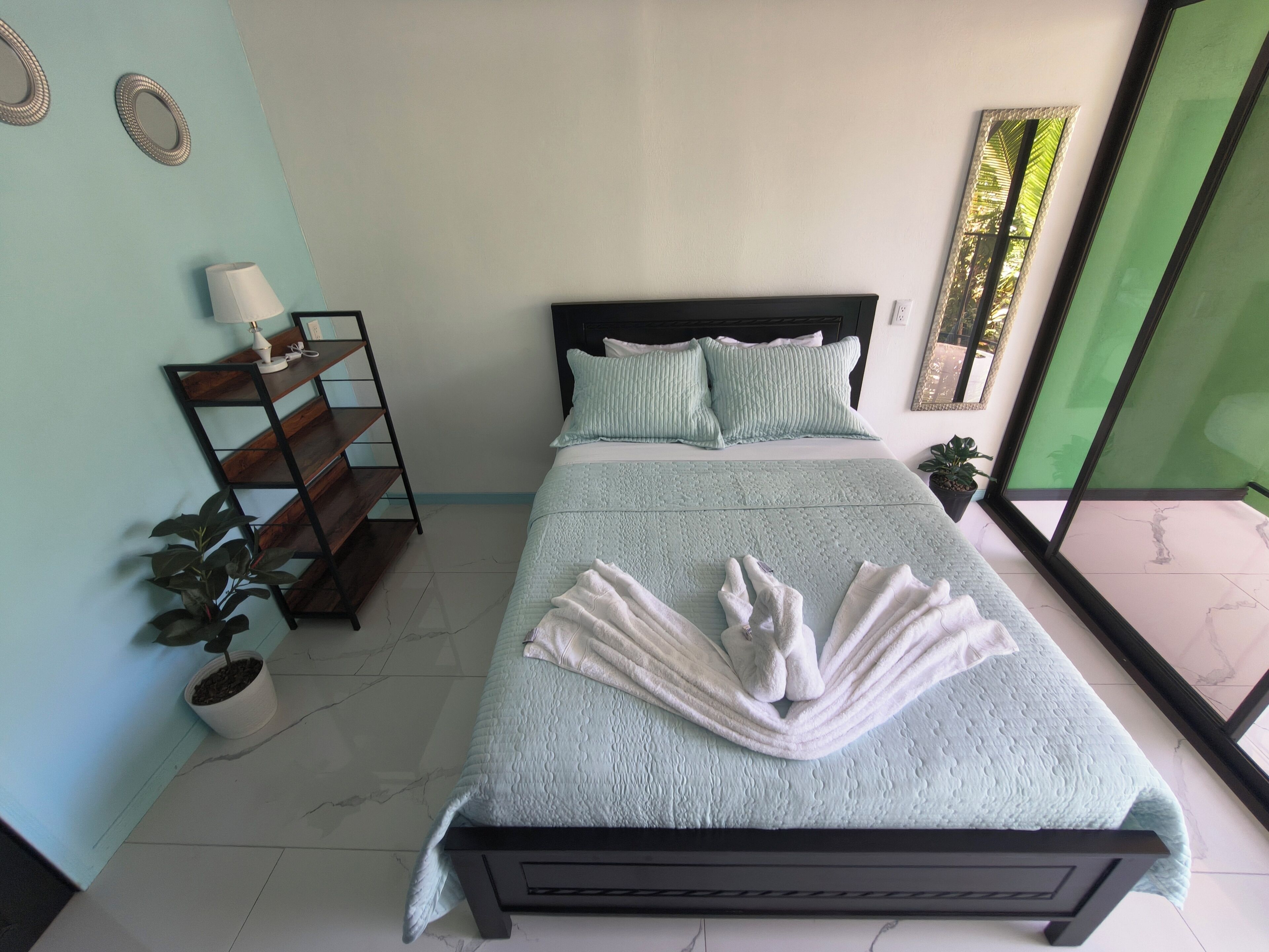 A bedroom with a bed, pillows, a bedside table with a lamp, a plant, and a view of the outside.