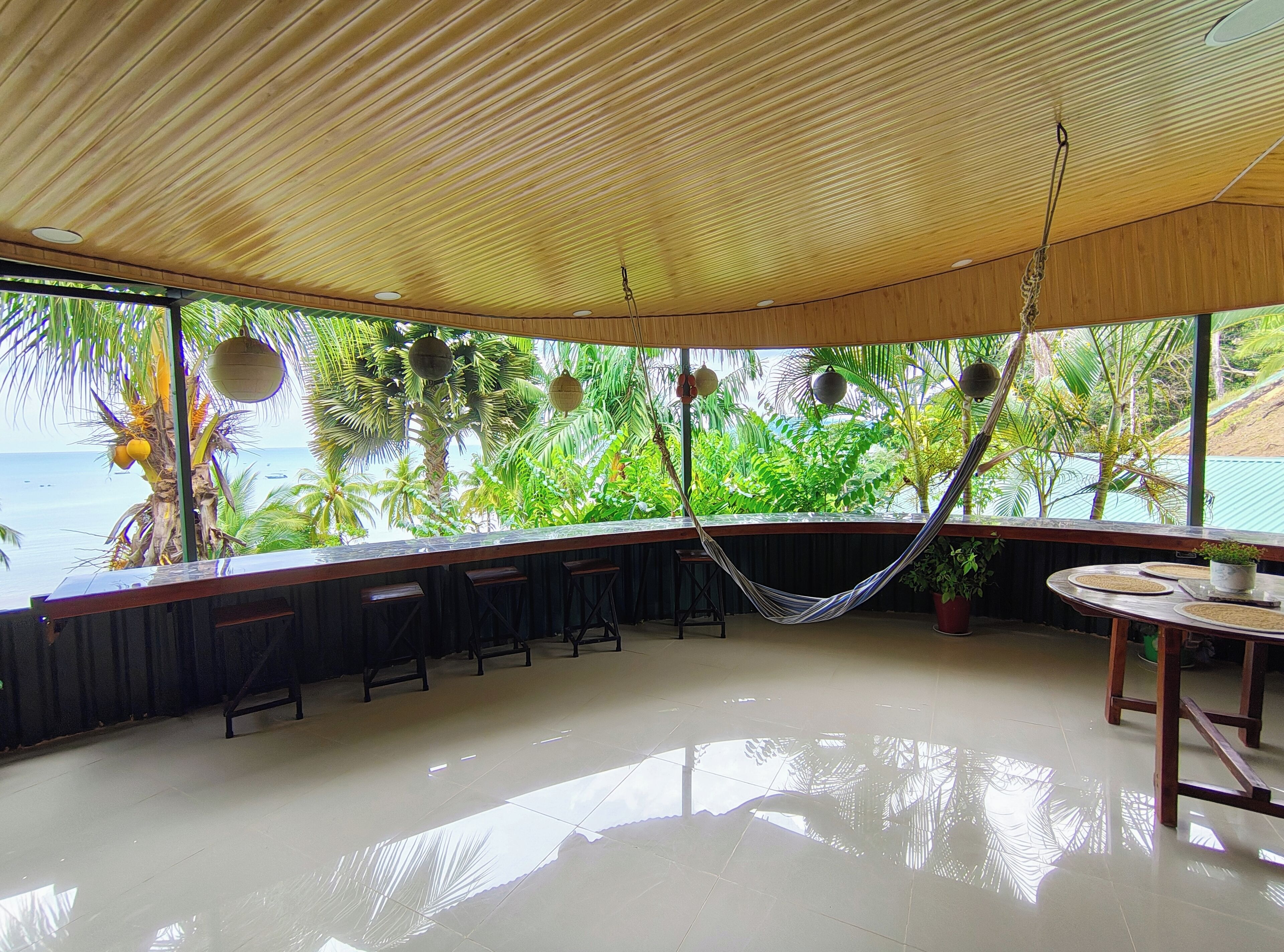 A spacious room with a wooden ceiling, a hammock, a bar area with stools, and a view of the ocean through large windows.