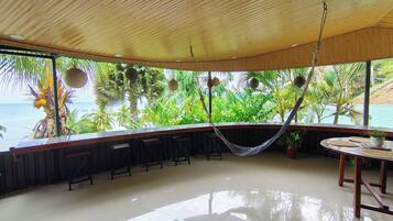 A spacious room with a wooden ceiling, a hammock, a bar area with stools, and a view of the ocean through large windows.