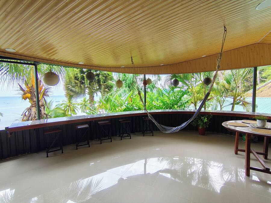 A spacious room with a wooden ceiling, a hammock, a bar area with stools, and a view of the ocean through large windows.