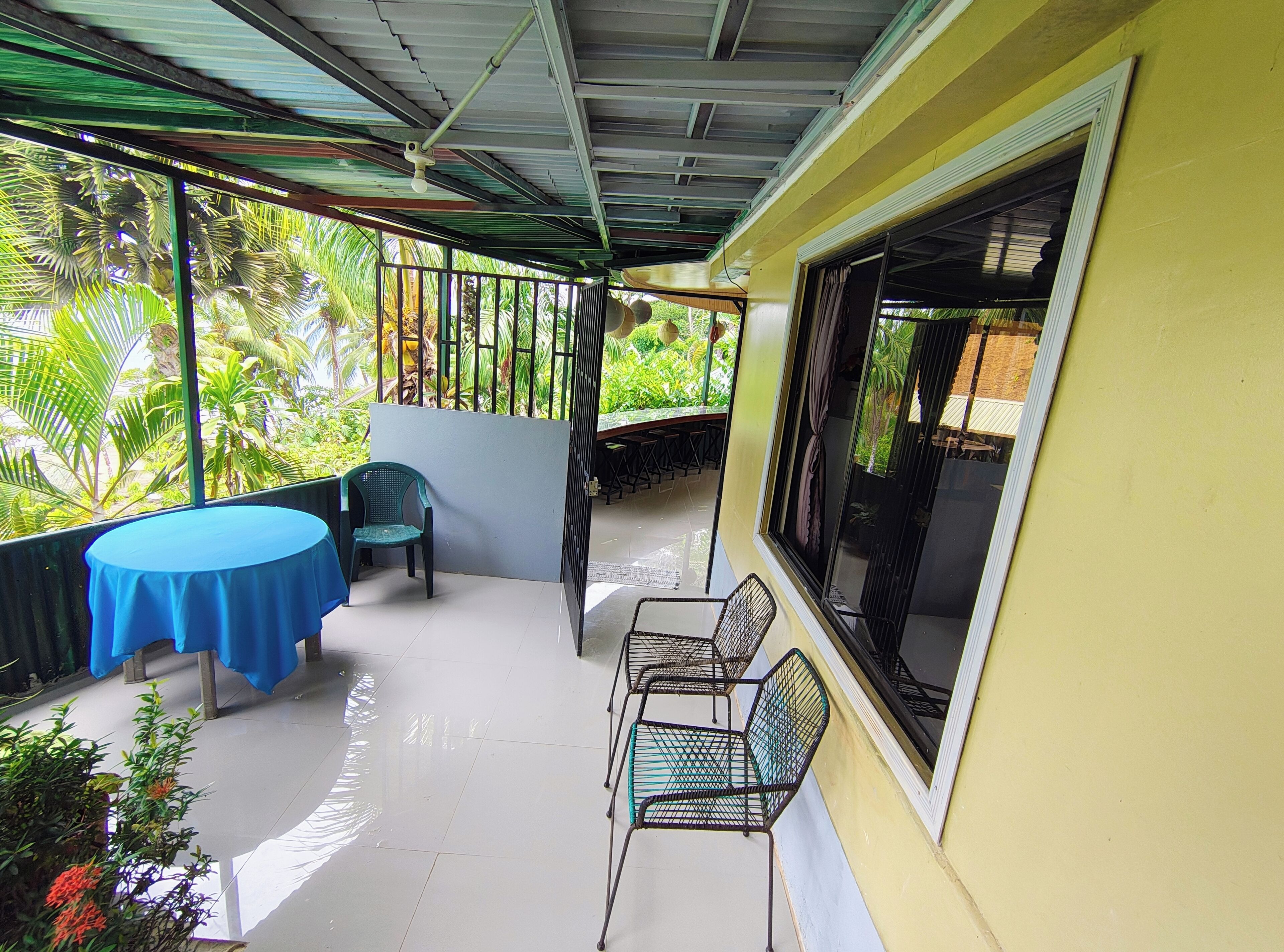 A covered outdoor area with a table and chairs, a window, and a view of greenery.