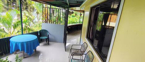 A covered outdoor area with a table and chairs, a window, and a view of greenery.