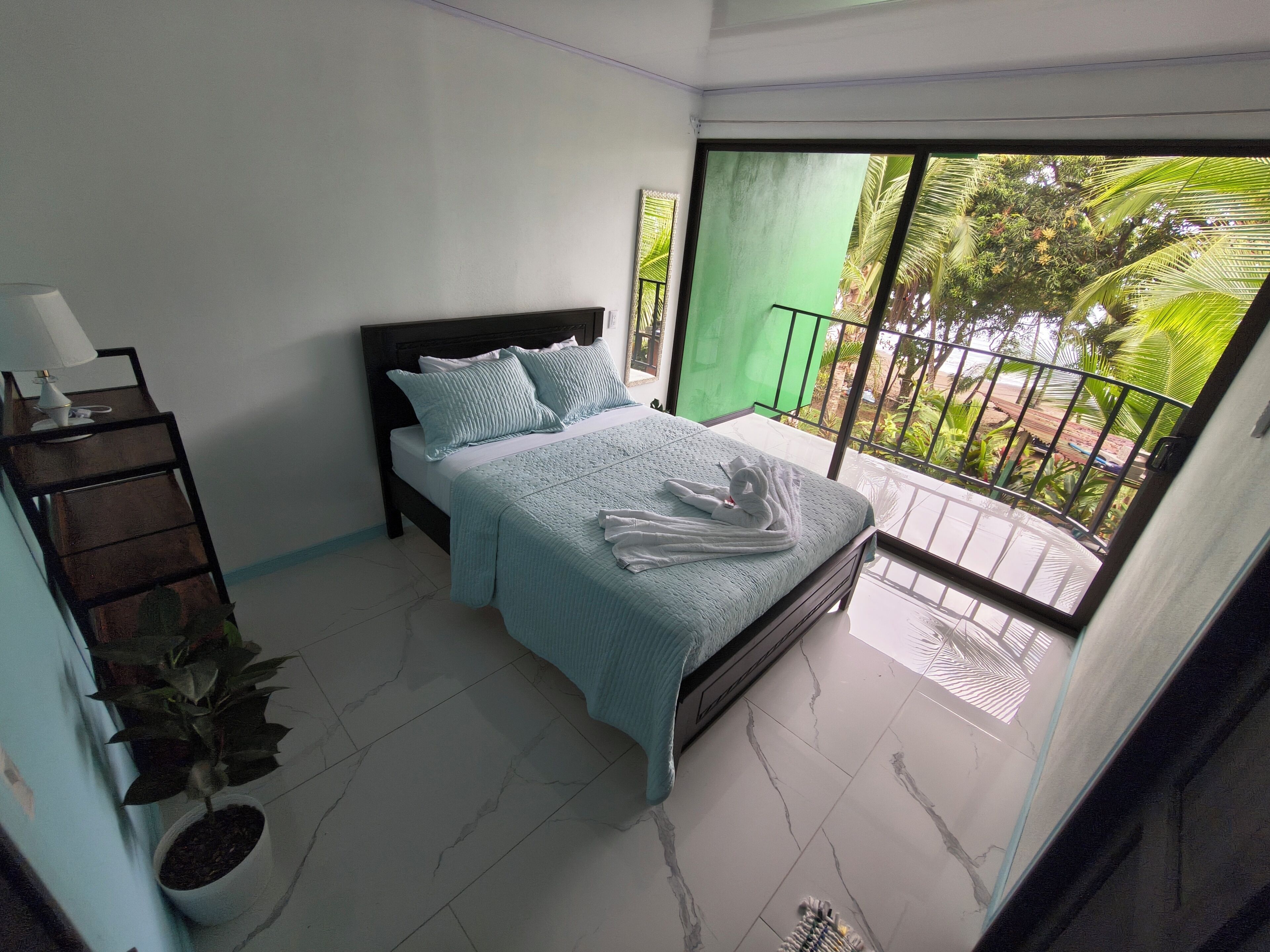 A bedroom with a bed, a nightstand, a potted plant, and a balcony with a view of greenery.