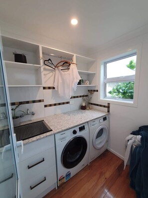 A compact laundry room with a washing machine, drying rack, and shelving. - Garden View, Sherwood Forest - Tauranga (Tauranga)