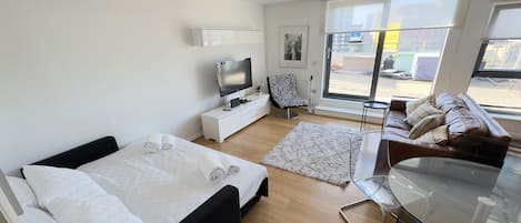 A modern hotel room with a bed, a TV, a sofa, a glass table, and a large window offering a view of the outside.