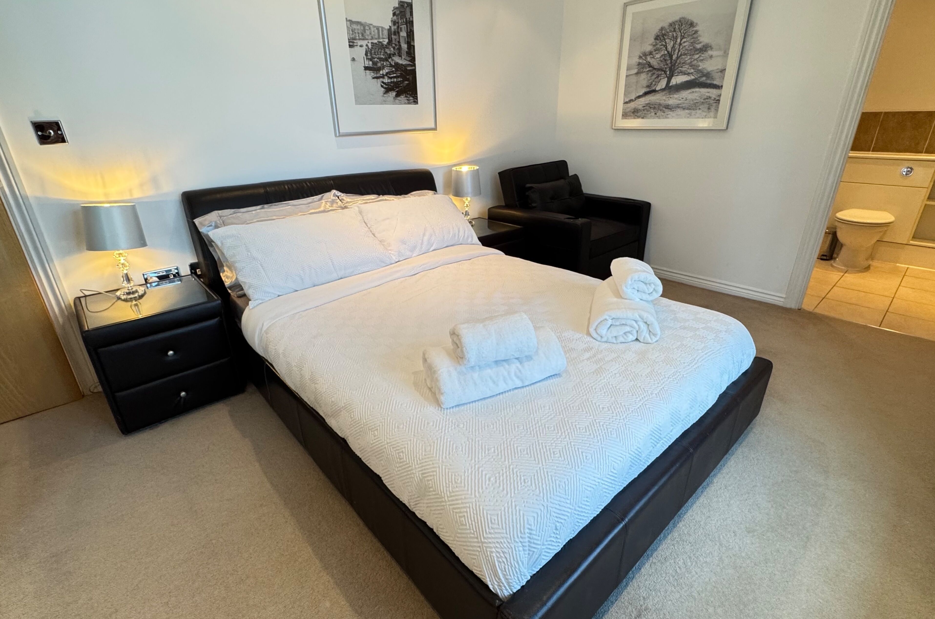 A neatly made bed with white linens and a folded towel, a black nightstand with a lamp, and a black chair.