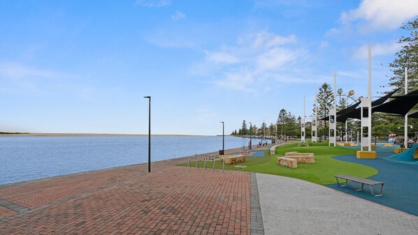 A waterfront park with a paved walkway, benches, and a playground area.