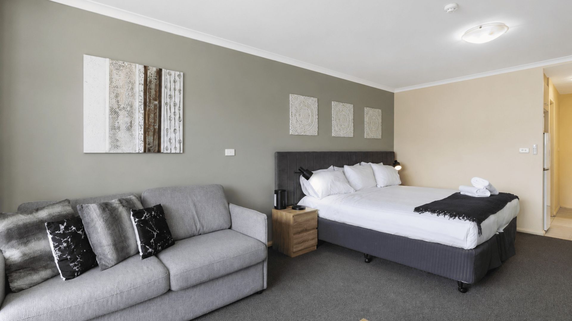 A modern hotel room with a grey sofa, a large bed with white bedding, and three wall-mounted artworks.