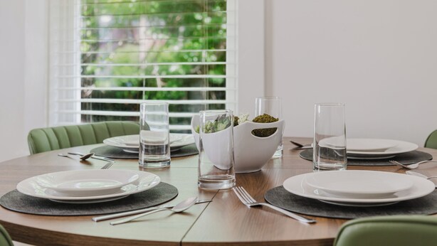 Una mesa de comedor dispuesta con platos, cubiertos y vasos, con una planta en maceta y una ventana con persianas al fondo.