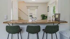 Una cocina con encimera de madera, tres sillas tapizadas en verde, un cubo de basura blanco y una escalera que conduce a otra habitación.