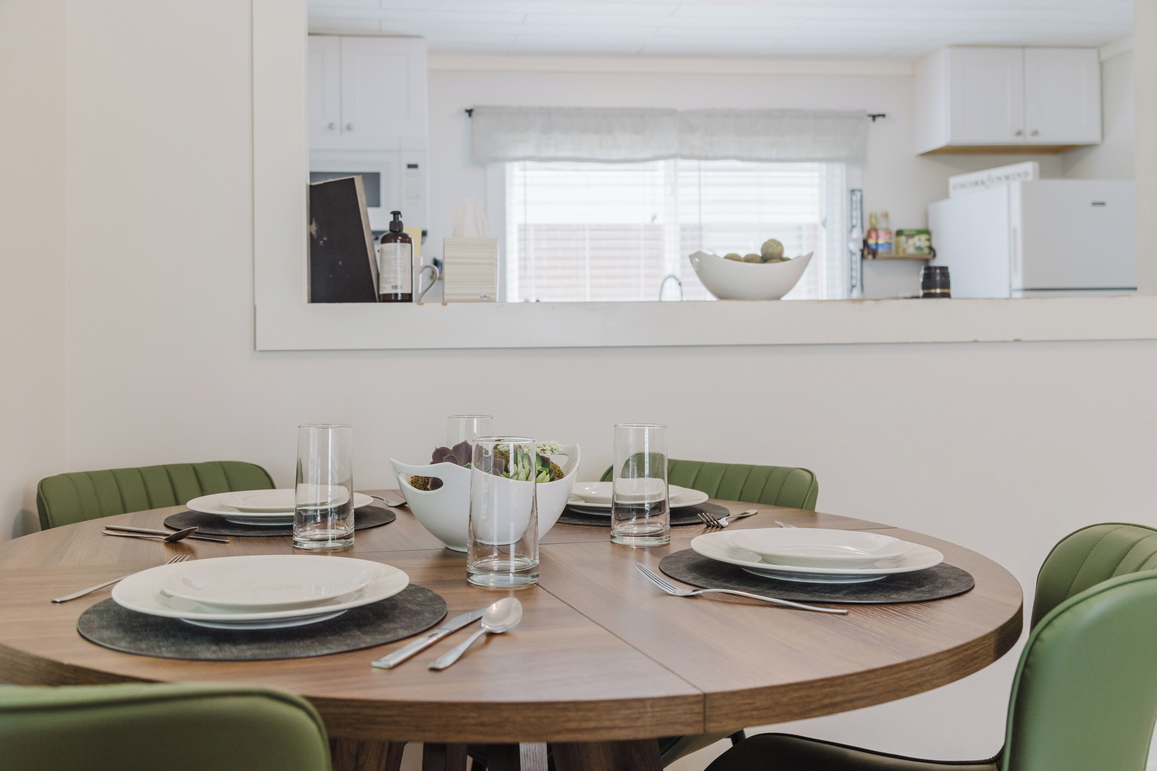 Une table à manger dressée avec des assiettes, des verres et des couverts, dans une cuisine moderne aux armoires blanches et avec un grand miroir au-dessus.