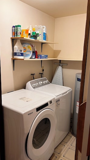 A laundry area with a Whirlpool washer and dryer, shelves with cleaning supplies, and a water heater.