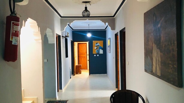 Un couloir avec un extincteur, un luminaire au plafond, une porte donnant sur une autre pièce et une chaise.