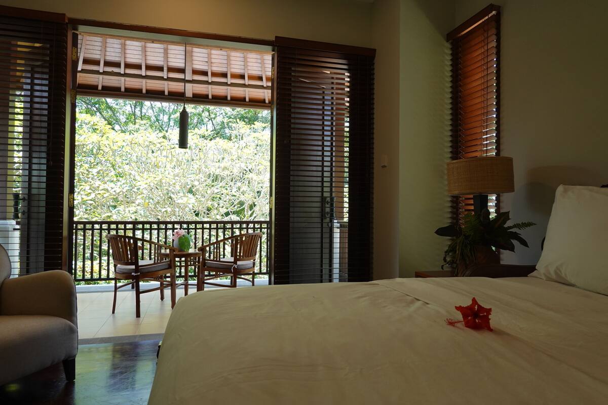 Habitación de hotel con cama, una mesa y sillas, y un balcón con vistas a zonas verdes.