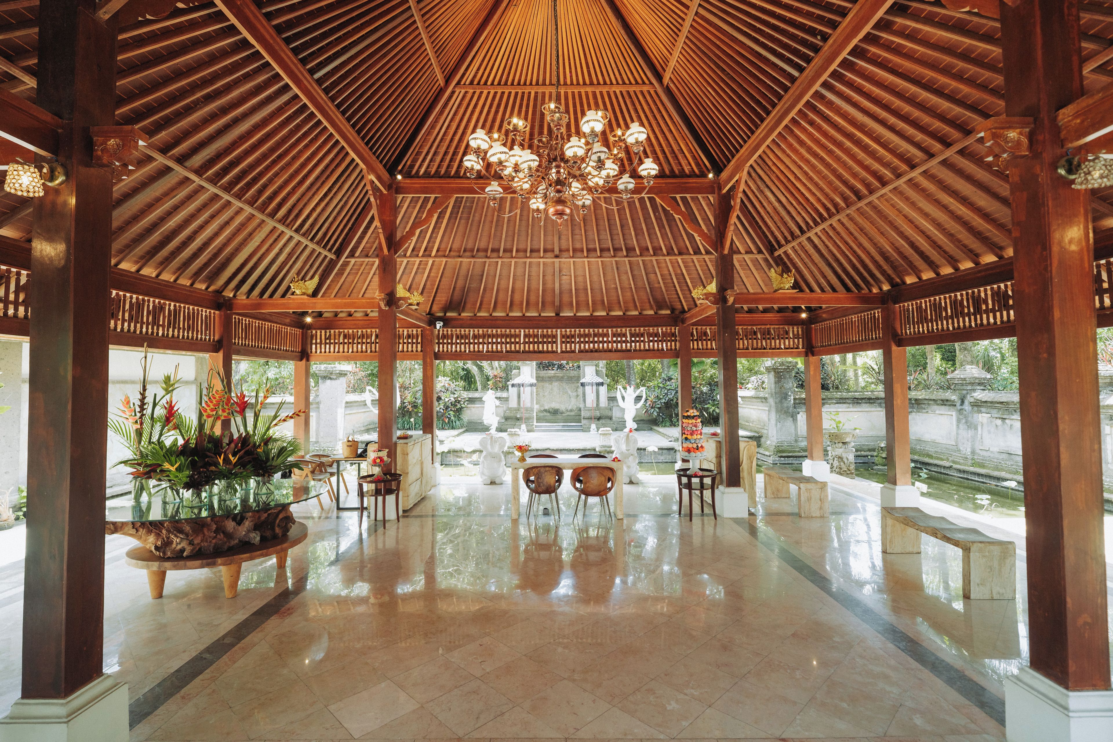 A spacious indoor area with a high ceiling and a central chandelier, featuring a glass table and chairs, and a view of an outdoor garden.