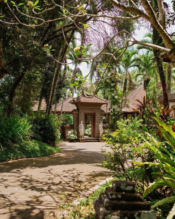 A pathway leading to a traditional building with a tiled roof, surrounded by lush greenery and palm trees.