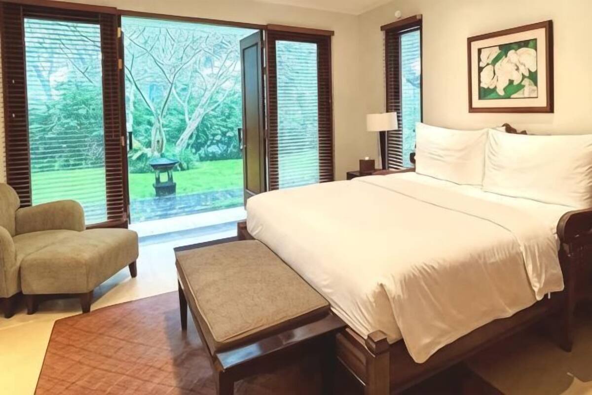 Un dormitorio con cama, una silla, un banco y vistas a un jardín a través de ventanas abiertas.