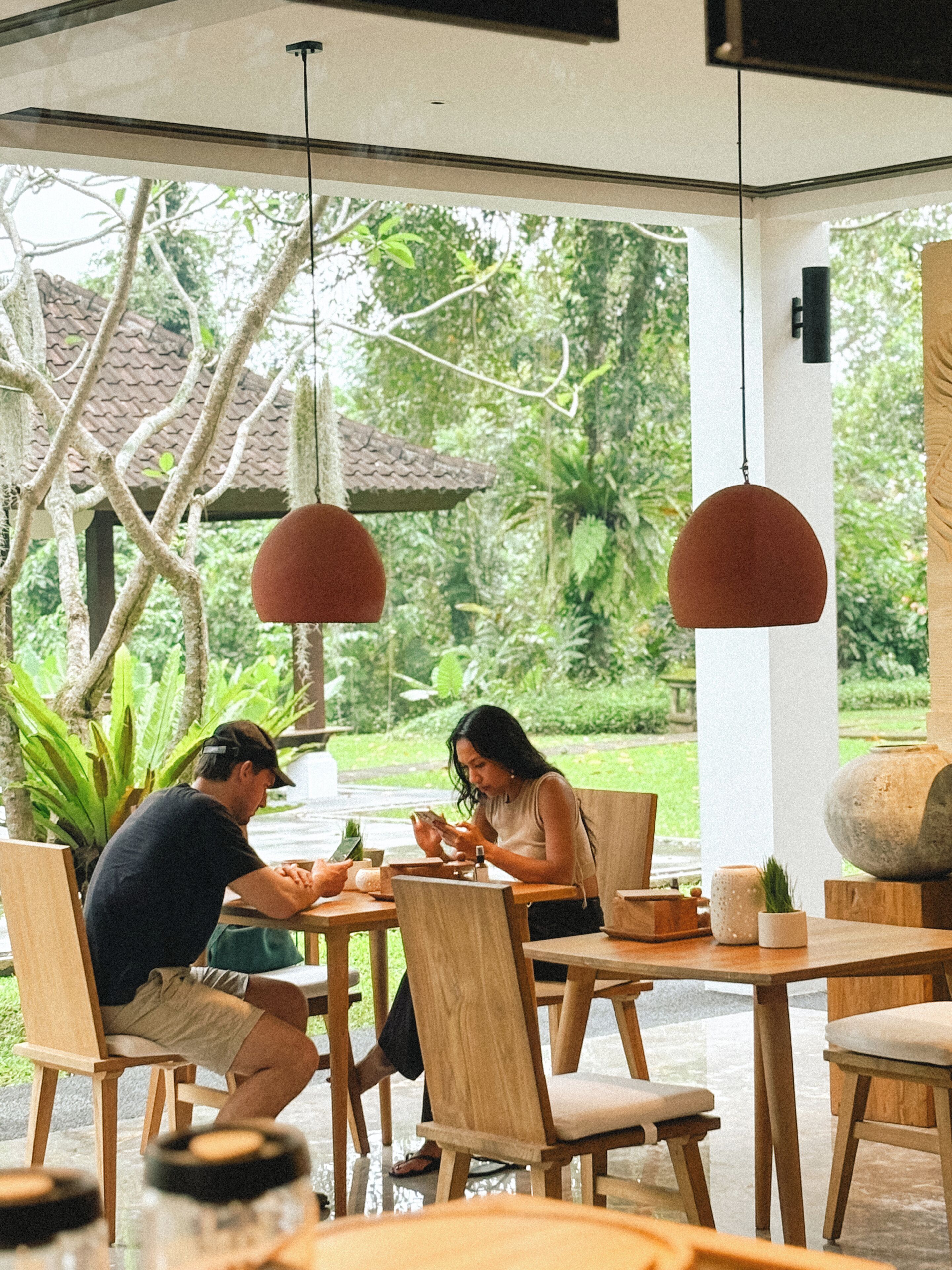An outdoor dining area with wooden tables and chairs, surrounded by greenery and a view of a traditional building.
