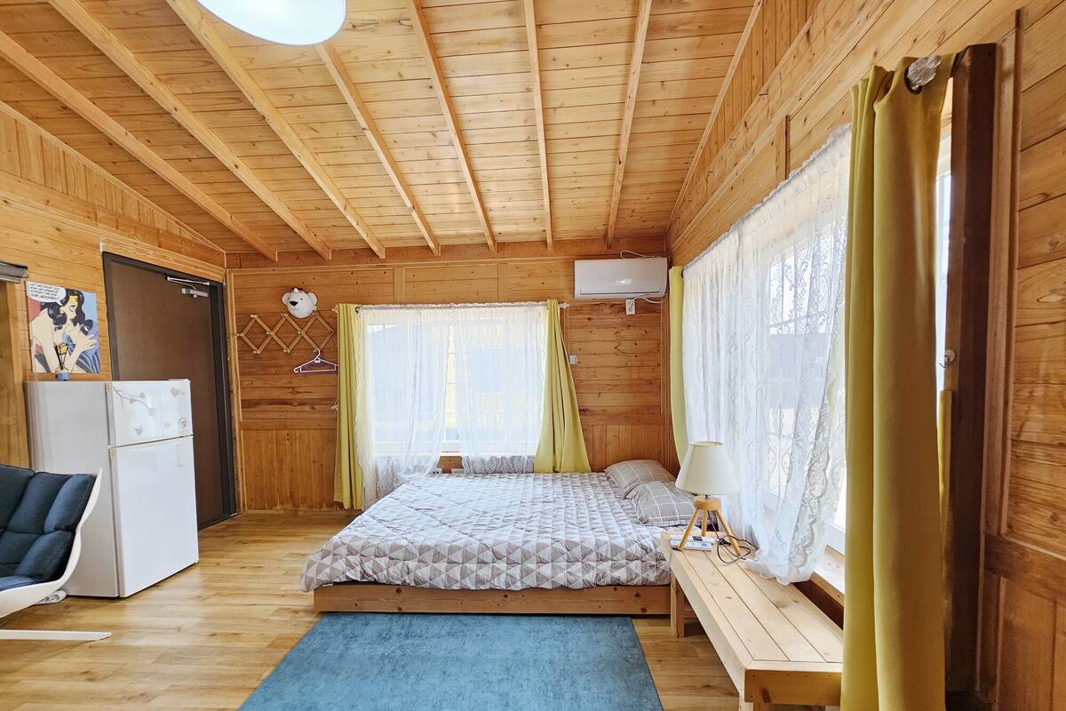 A cozy wooden cabin room with a bed, a desk, a chair, and a refrigerator.