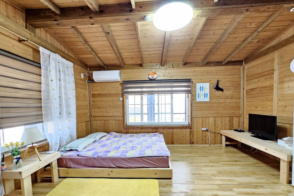 A wooden cabin room with a bed, a desk, a TV, and a window with blinds.