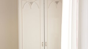 A white wardrobe with two doors and a lower shelf, a wooden bed with a white mattress, and a white curtain partially visible.