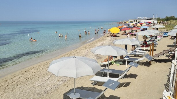 A beach with sun loungers and umbrellas, a building labeled 'BIO MANIA' in the background, and people swimming in the water.