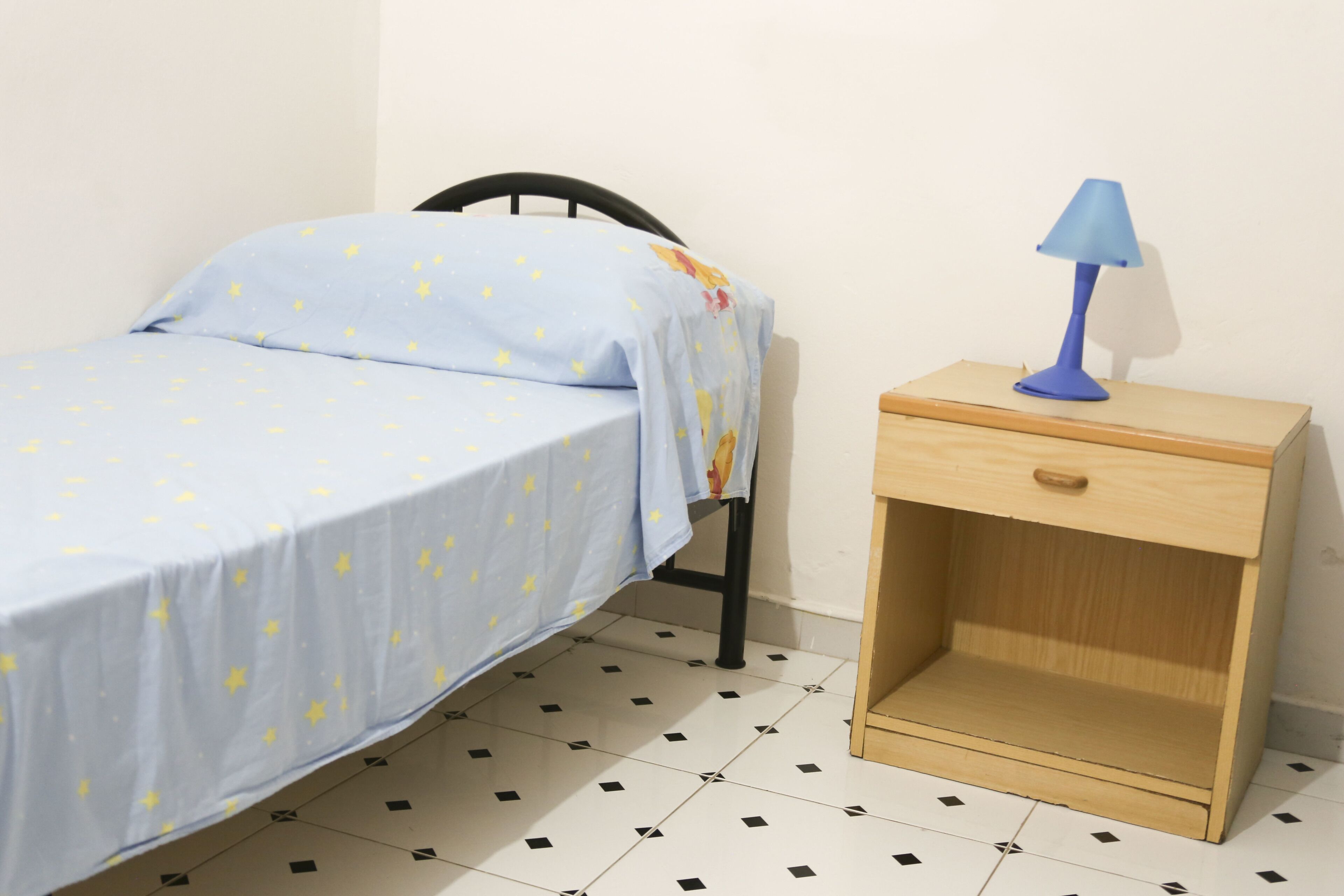Una cama individual con colcha azul y una mesita de noche pequeña con lámpara.