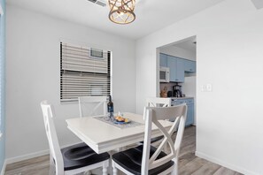 Un comedor con una mesa y sillas blancas, una ventana con persianas, y una cocina al fondo.