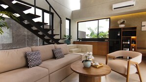 Una sala moderna con un sofá, una mesita de centro y una silla. Se ve una escalera y una cocina al fondo.