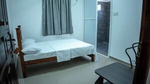 Una habitación pequeña con cama individual, estructura de cama de madera, colchón blanco y cortina gris. Hay una mesita y una silla en la esquina, y una puerta que da a una zona con baldosas.