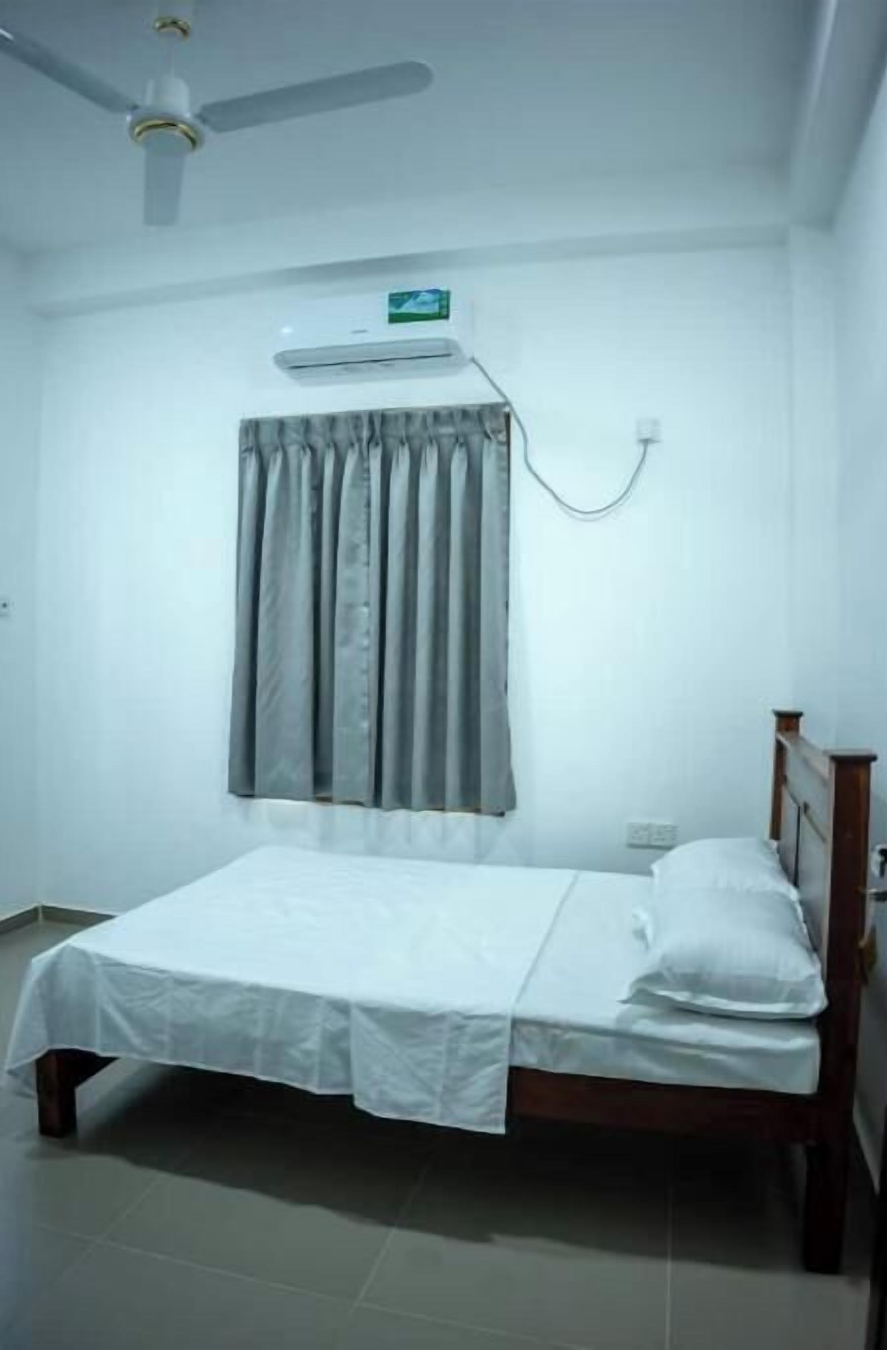 A single-bed room with a ceiling fan, an air conditioning unit, and a window with grey curtains.