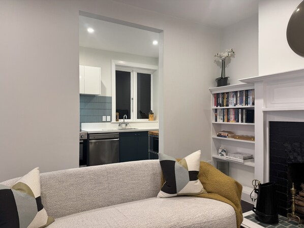 Un salon moderne avec un canapé, une cheminée, une bibliothèque et une cuisine visible à travers une porte ouverte.