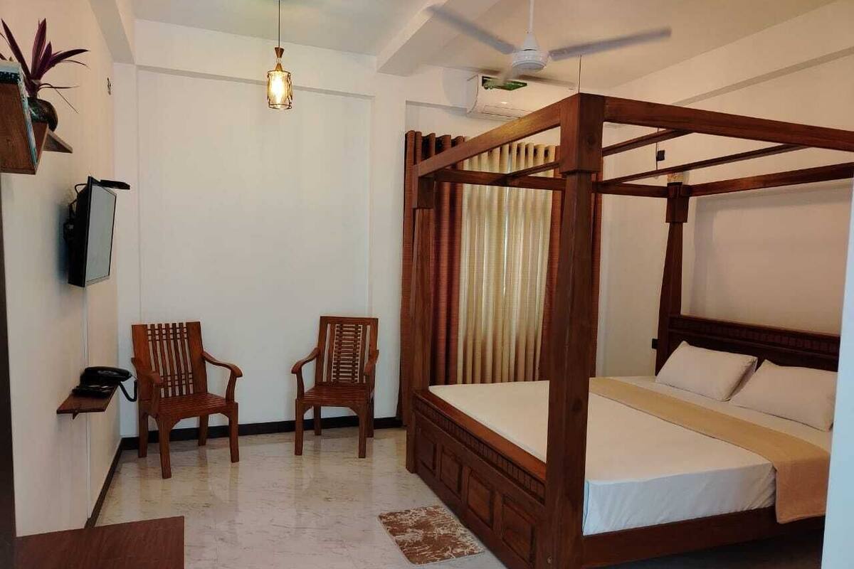 Una cama con dosel de madera, ropa de cama blanca, dos sillas de madera, una mesita con un teléfono y un ventilador de techo.