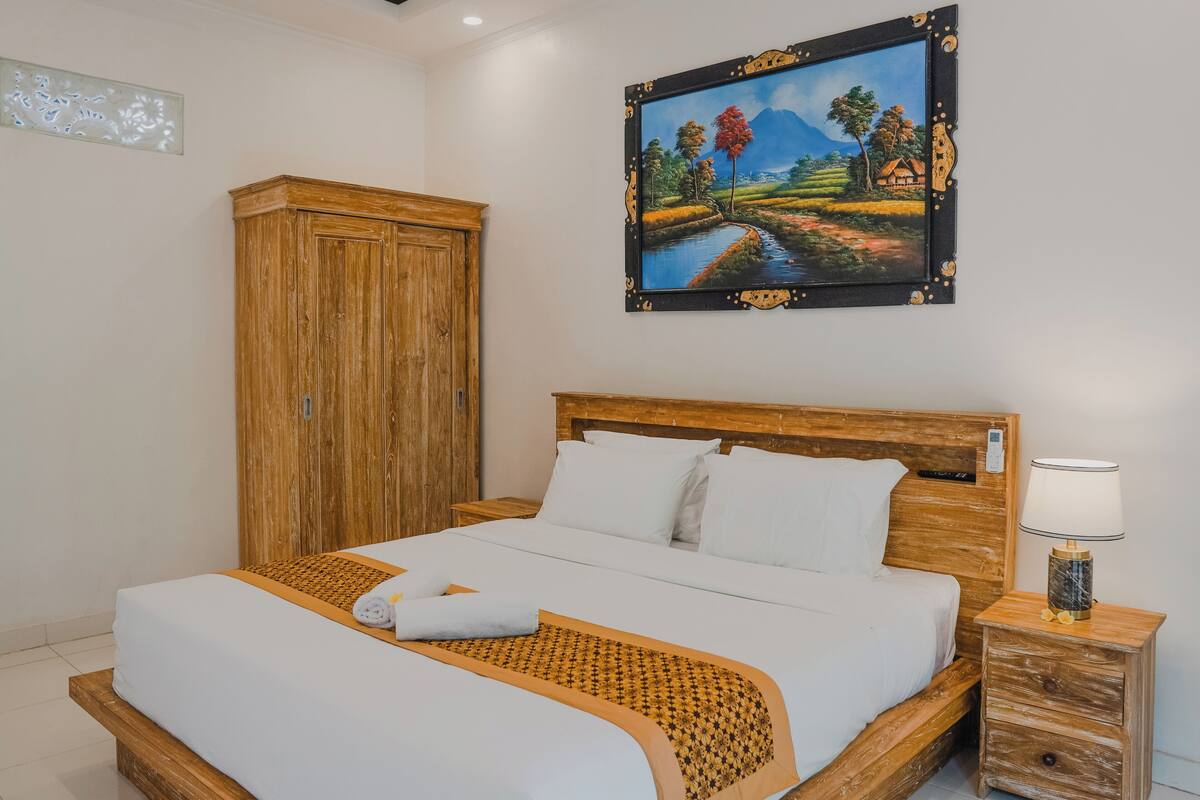 Een net opgemaakt bed met witte lakens en een decoratieve plaid, een houten garderobe, een nachtkastje met een lamp en een landschapsschilderij aan de muur.