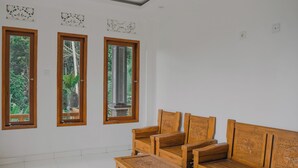 Een kamer met een houten bank en stoelen, grote ramen met uitzicht op groen, en decoratieve wandpanelen.