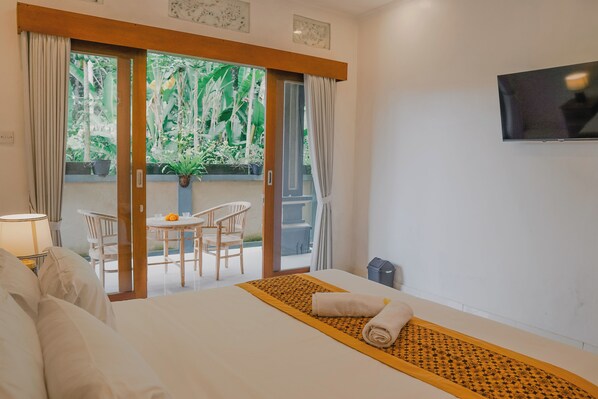 Habitación de hotel con cama, televisor, ventana con cortinas, una pequeña mesa con sillas y vistas a zonas verdes.