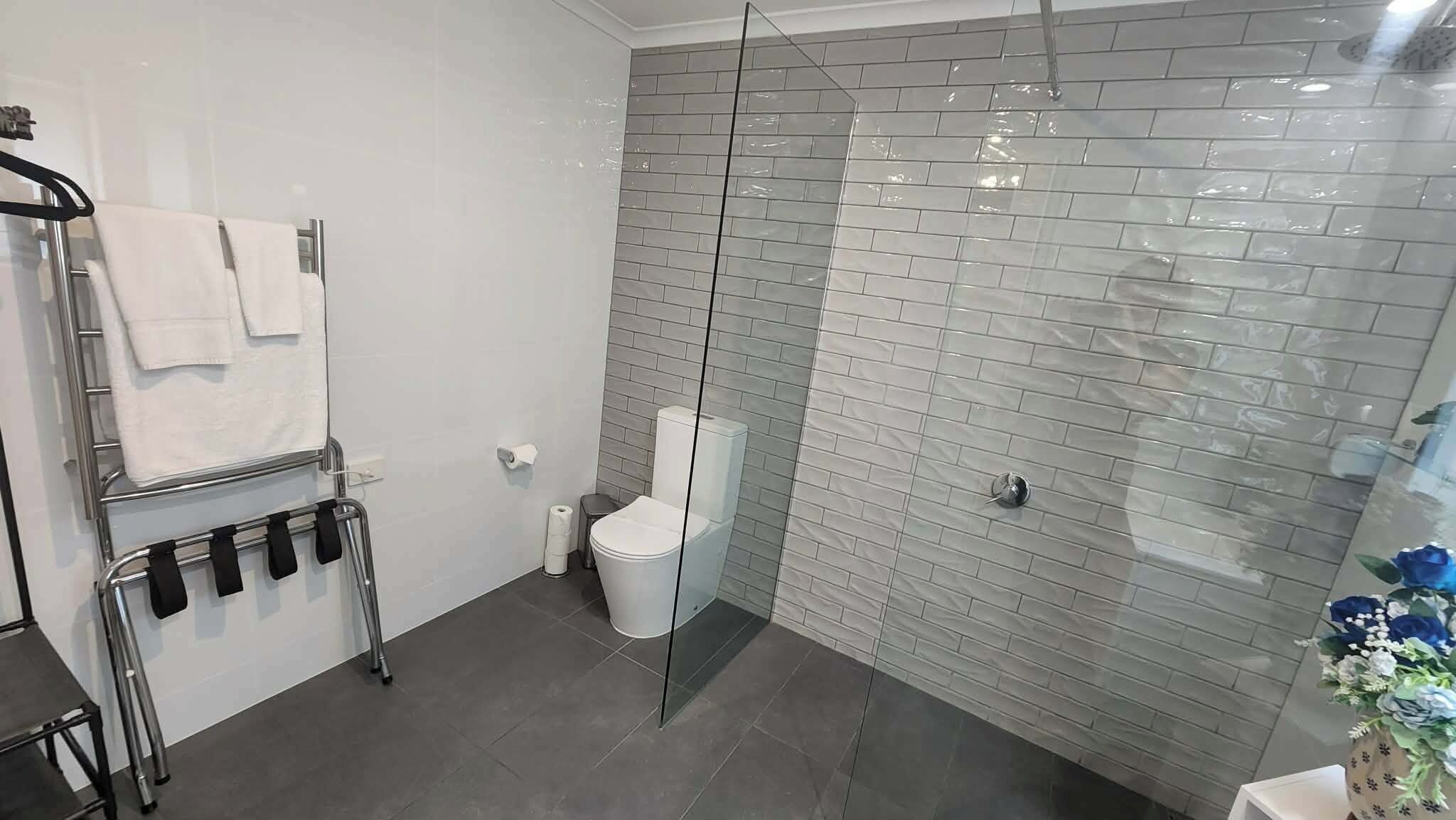 A bathroom with a glass shower enclosure, a towel rack with towels, a toilet, and a small trash bin.