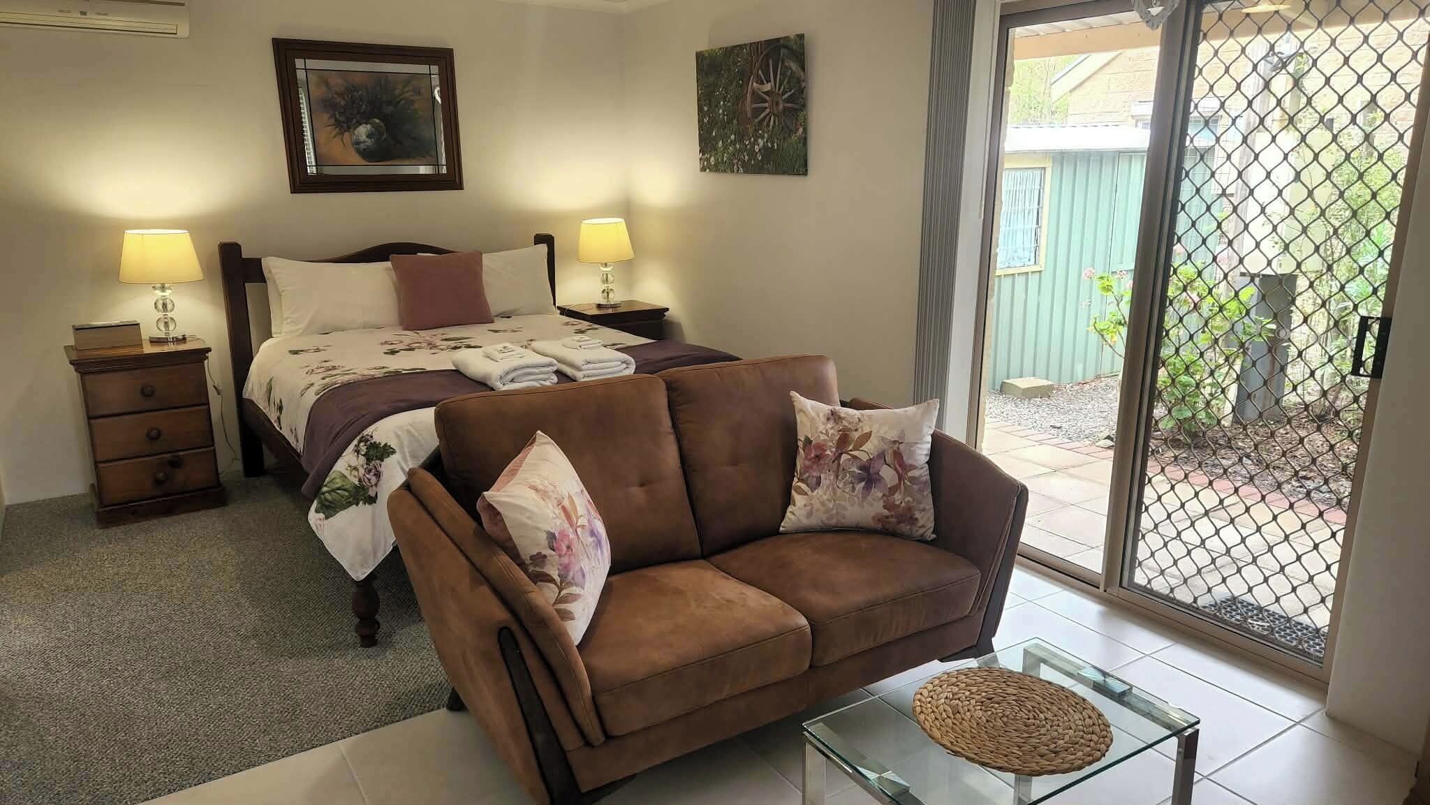 A bedroom with a bed, two bedside tables with lamps, a brown sofa, a glass coffee table, and a view through a sliding glass door.